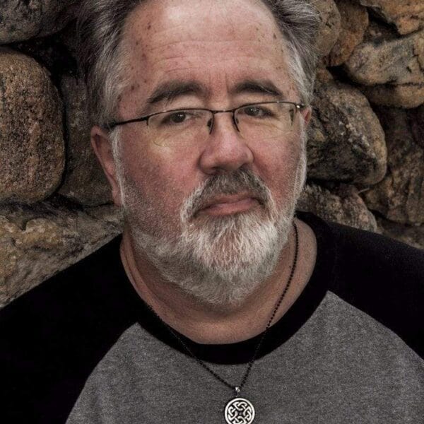 A man with glasses and a beard leaning against a stone wall.
