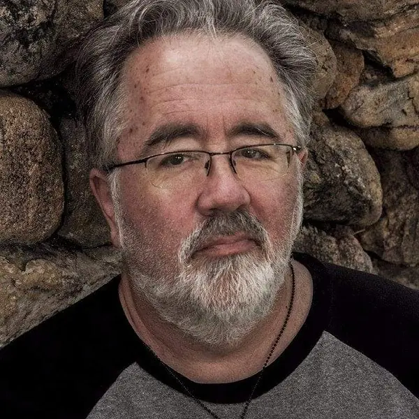 A man with glasses and a beard leaning against a stone wall.