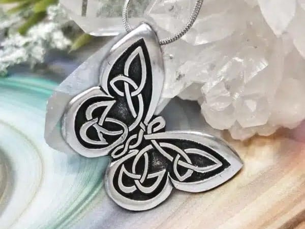 A silver pendant with Celtic knot motifs arranged in a butterfly shape, placed next to a cluster of clear crystals on a multicolored surface.