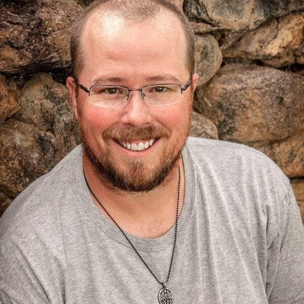 A man with glasses smiling in front of a stone wall.