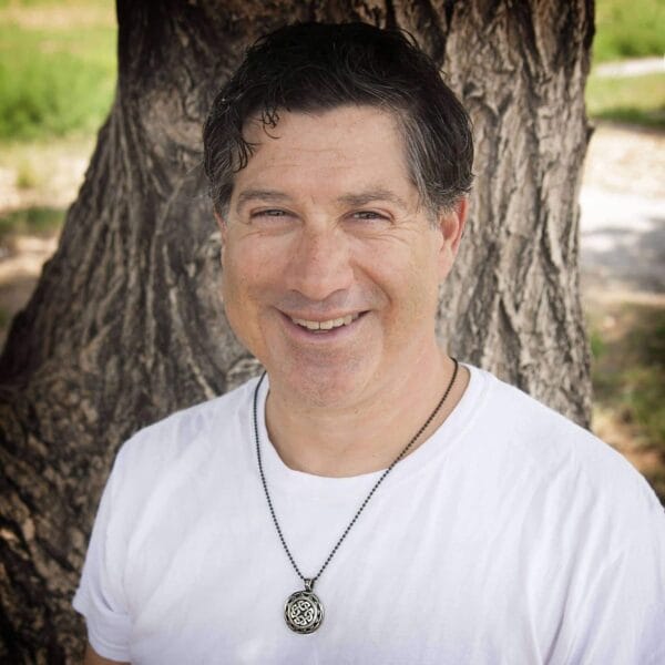A man in a white t - shirt standing next to a tree.