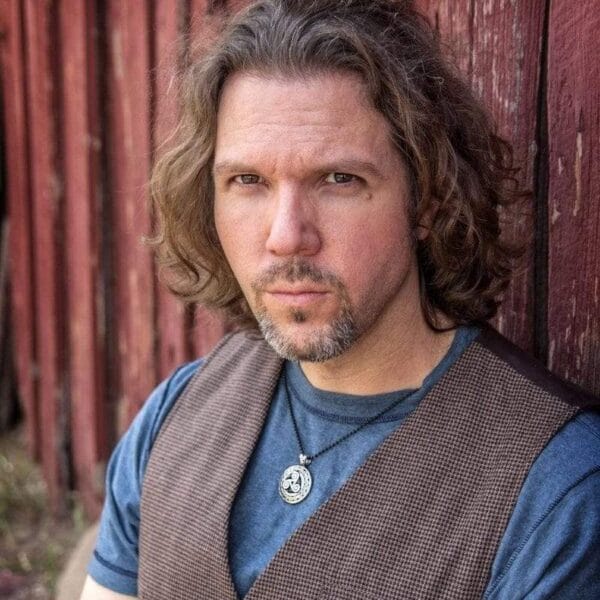 A man with long hair leaning against a red barn.