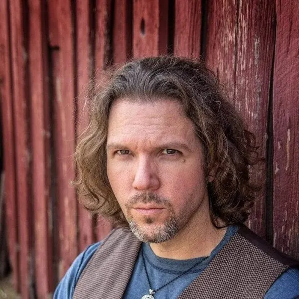 A man with long hair leaning against a red barn. A man with long hair leaning against a red barn.