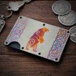 A Celtic Bear Titanium Minimalist Wallet featuring a colorful bear design and intricate Celtic knot patterns is placed on a wooden surface next to several metallic keychains and coins with detailed designs.