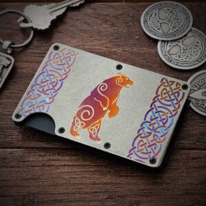 A Celtic Bear Titanium Minimalist Wallet featuring a colorful bear design and intricate Celtic knot patterns is placed on a wooden surface next to several metallic keychains and coins with detailed designs.
