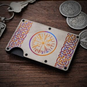 The Wayfinder Titanium Minimalist Wallet, featuring vibrant Norse designs and runes, is placed on a wooden surface near keychains and coins.
