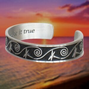 A Dragonfly Dreams Bracelet, featuring intricate dragonfly and spiral designs, is displayed against a sunset background over a beach. The inner side reads, "Make it true.