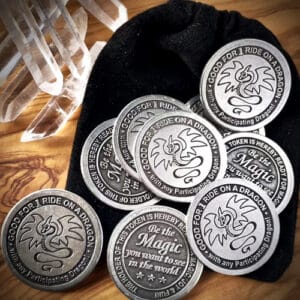 A collection of Magical Dragon Ride Tokens 10 Pack!, each featuring a dragon insignia and the text "GOOD FOR 1 RIDE ON A DRAGON" alongside "Be the Magic you want to see in the world," are arranged on a wooden surface beside a black pouch.