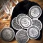 A collection of Magical Dragon Ride Tokens 10 Pack!, each featuring a dragon insignia and the text "GOOD FOR 1 RIDE ON A DRAGON" alongside "Be the Magic you want to see in the world," are arranged on a wooden surface beside a black pouch.