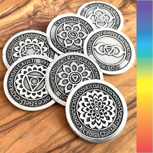 Six coins from the Chakra Blessing 7 pc Oracle Set, each adorned with chakra designs and inscriptions, are arranged on a wooden surface. A vertical strip with rainbow colors is visible on the right.