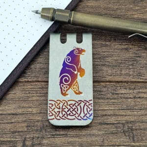 A metal bookmark with a colorful bear design and Celtic patterns, placed on a wooden surface next to a pen and dotted notebook.