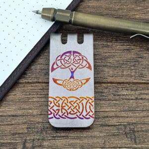 Metal bookmark with intricate Celtic knot designs next to a pen and notebook on a wooden surface.