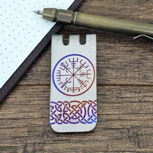 Metal bookmark with a Viking compass design and intricate knot pattern, placed on a wooden surface next to a bronze pen and a dotted notebook.