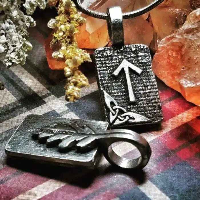 Three metal pendants with Viking style designs, including an arrow, a feather, and a triquetra, are displayed on a checkered fabric with crystal accents and dried flowers.