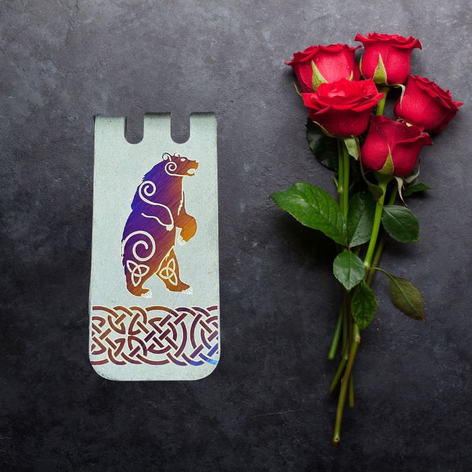 A bookmark with a colorful bear design lies next to a bouquet of five red roses on a dark surface.