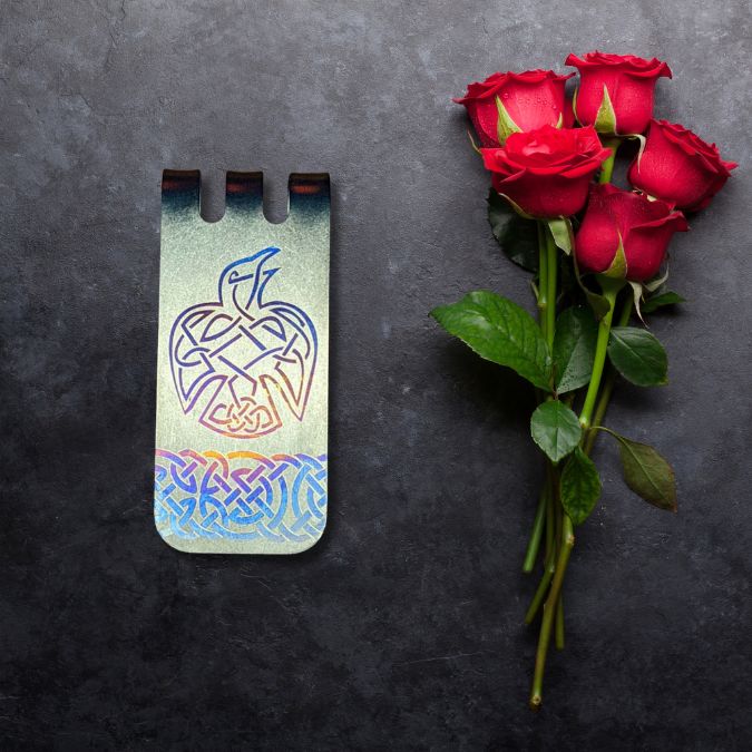 A bouquet of red roses lies next to a decorative metal bookmark with a Celtic design depicting a bird, on a dark surface.