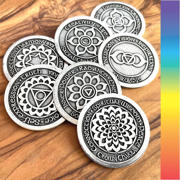 Seven silver coins with chakra symbols and text on a wooden surface, next to a vertical rainbow stripe.