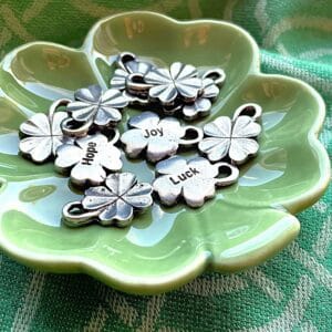 The Bits of Luck Holiday Limited Edition features silver clover shaped charms with inscriptions such as 'Hope,' 'Joy,' and 'Luck,' perfectly displayed in a green clover shaped dish.
