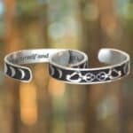 Two metal bracelets with engraved designs: the Triple Moon Bracelet with a moon phase pattern and another featuring a floral pattern, against a blurred nature background.