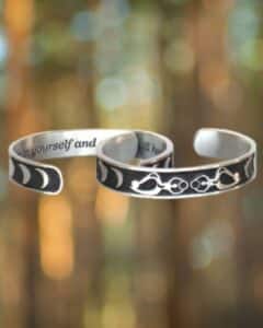 Two metal bracelets with engraved designs: the Triple Moon Bracelet with a moon phase pattern and another featuring a floral pattern, against a blurred nature background.