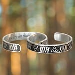 Two Sisterhood Bracelets in silver feature engraved designs of figures and symbols displayed against a blurred forest background.