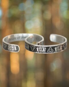 Two Sisterhood Bracelets in silver feature engraved designs of figures and symbols displayed against a blurred forest background.