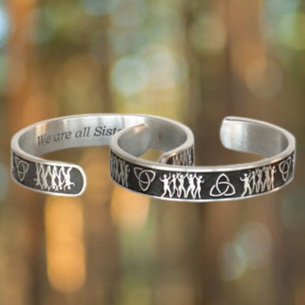 Two Sisterhood Bracelets in silver feature engraved designs of figures and symbols displayed against a blurred forest background.