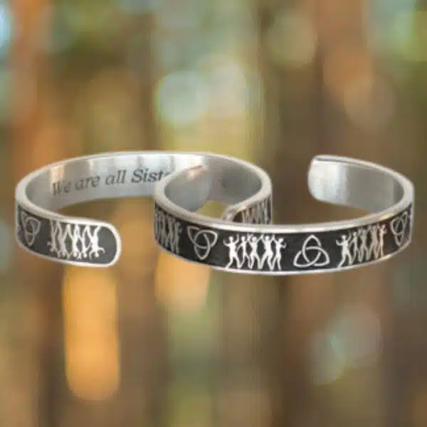 Two Sisterhood Bracelets in silver feature engraved designs of figures and symbols displayed against a blurred forest background.