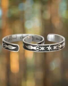Two Triple Aspect Goddess Bracelets, featuring silver open cuffs with engraved abstract designs and text on their inner surfaces, are set against a blurred background with circular light spots.