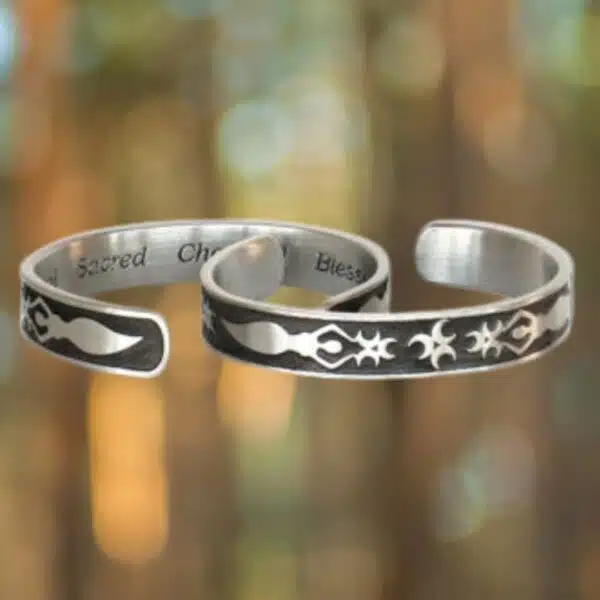 Two Triple Aspect Goddess Bracelets, featuring silver open cuffs with engraved abstract designs and text on their inner surfaces, are set against a blurred background with circular light spots.