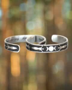 The Triple Moon Bracelet set includes two silver metal cuffs with celestial designs of moons and stars, highlighted against a blurred forest backdrop.