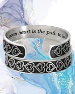 The Eve's Heart Bracelet, a pewter cuff featuring engraved heart motifs and the phrase "An open heart is the path to love," is displayed on an abstract blue and gold backdrop.