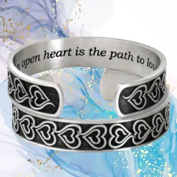 The Eve's Heart Bracelet, a pewter cuff featuring engraved heart motifs and the phrase "An open heart is the path to love," is displayed on an abstract blue and gold backdrop.