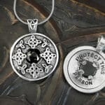 Two silver pendants with intricate designs are displayed. One is engraved with "PROTECT THIS PERSON" surrounding a smoky quartz, part of the limited edition Protection Pendant collection. Both rest on a textured surface.