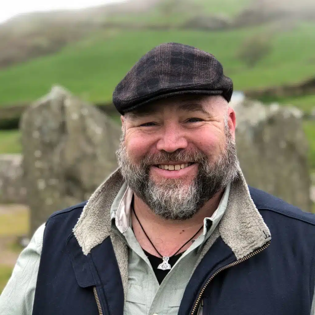 A bearded man wearing a flat cap and a jacket stands outdoors, smiling, with grassy hills and large stones in the background.