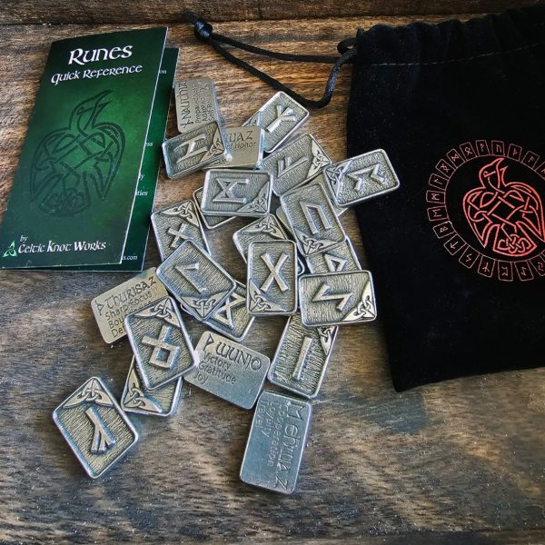 A set of metal runes is spread on a wooden surface next to a black drawstring pouch and a runes quick reference guide.