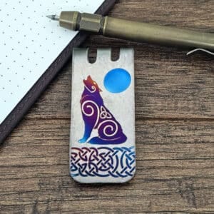 A titanium wolf money clip with Celtic knot and swirl designs, featuring a stylized howling wolf against a blue moon, displayed on a wooden surface beside a notepad and pen.