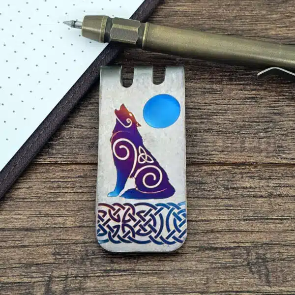 A titanium wolf money clip with Celtic knot and swirl designs, featuring a stylized howling wolf against a blue moon, displayed on a wooden surface beside a notepad and pen.