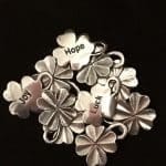 A cluster of silver four leaf clover charms with the words "Hope," "Luck," and "Joy" engraved on three of them, displayed against a black background.
