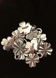 A cluster of silver four leaf clover charms with the words "Hope," "Luck," and "Joy" engraved on three of them, displayed against a black background.