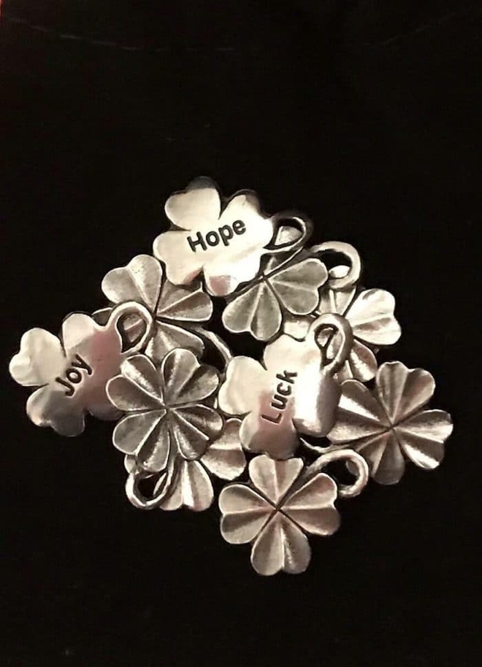 A cluster of silver four leaf clover charms with the words "Hope," "Luck," and "Joy" engraved on three of them, displayed against a black background.
