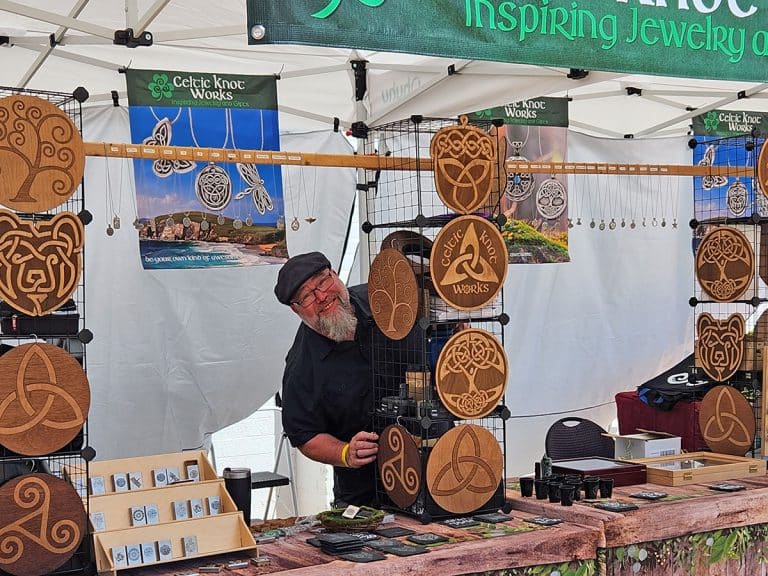 A vendor stands at a booth displaying wooden Celtic knot designs, jewelry, and art under a tent at an outdoor event.