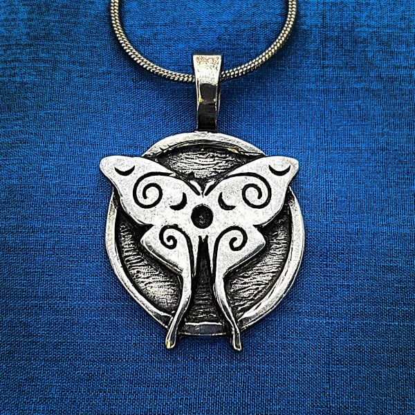 The Luna Moth Pendant, featuring swirling designs, hangs from a chain against a blue fabric background.