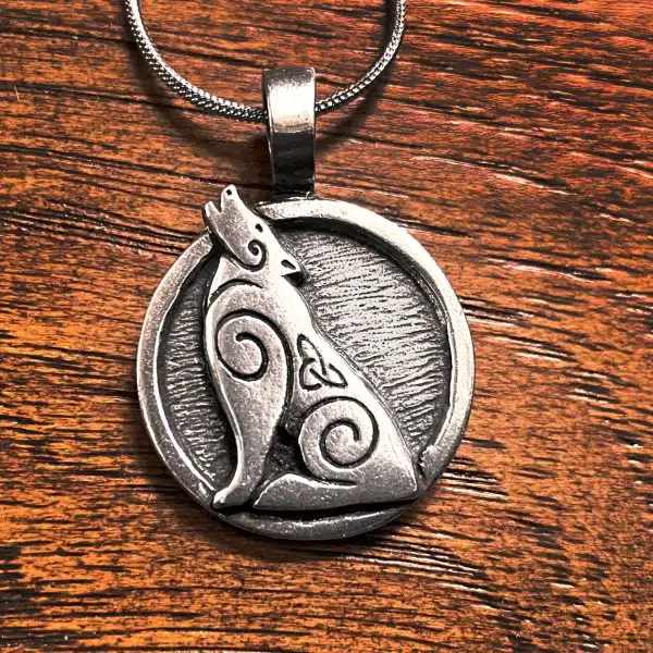 Howling Wolf Pendant The Howling Wolf Pendant is a round silver piece featuring a stylized howling wolf and Celtic inspired spiral patterns, displayed on a wooden surface.