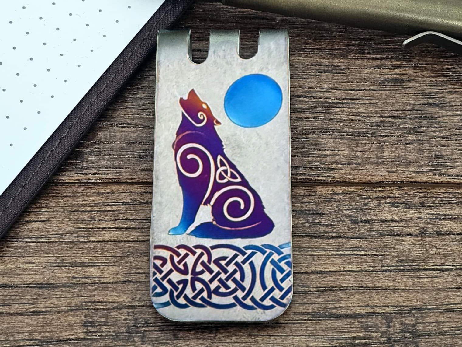 A metal money clip featuring a stylized, colorful wolf with Celtic knot designs, howling at a blue moon, set on a wooden surface.