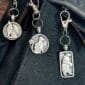 The Howling Wolf Bag Charm from the Celtic Spirals Collection features intricate swirling patterns, adding a unique touch to any dark fabric bag.