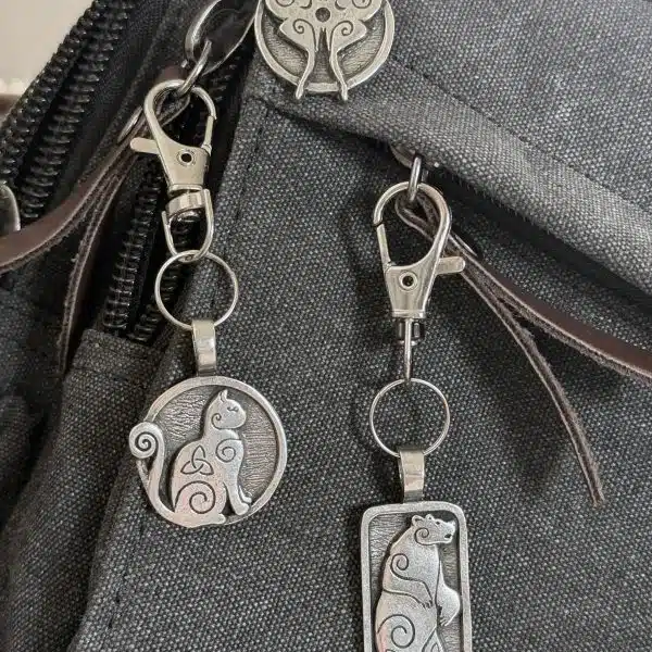 Luna Moth Bag Charm – Celtic Spirals Collection Three silver cat keychains and a Luna Moth Bag Charm Celtic Spirals Collection are attached to a black fabric bag. Each cat keychain features a unique shapeโcircle, rectangle, or cat face with swirling patternsโfor whimsical style.