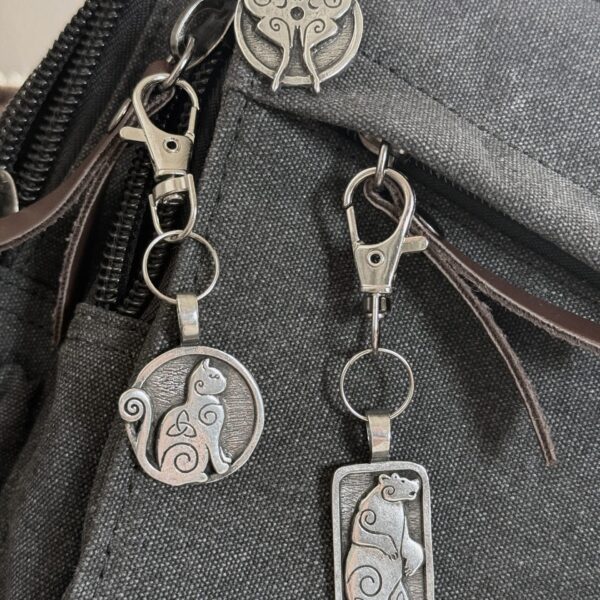 Luna Moth Bag Charm – Celtic Spirals Collection Three silver cat keychains and a Luna Moth Bag Charm Celtic Spirals Collection are attached to a black fabric bag. Each cat keychain features a unique shapeโcircle, rectangle, or cat face with swirling patternsโfor whimsical style.