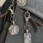 Three silver cat keychains and a Luna Moth Bag Charm Celtic Spirals Collection are attached to a black fabric bag. Each cat keychain features a unique shape—circle, rectangle, or cat face with swirling patterns—for whimsical style.