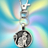 The Celtic Cat Bag Charm from the Celtic Spirals Collection features a silver keychain with a round pendant showing a stylized cat and Celtic swirls on a blue yellow tie dye background—perfect for bags or as a cat themed accessory.